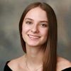 A young woman with long straight brown hair smiles softly against a blurred neutral background.