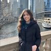 A young woman in a black coat smiles while standing on a stone bridge over a river with city buildings behind her.