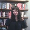 A young woman with dark hair and bangs smiles slightly while standing in front of bookshelves filled with various books.