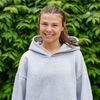 A young woman with brown hair tied back smiles at the camera wearing a light gray hoodie against a leafy green background.