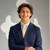 A young man with brown hair smiles wearing a dark pinstripe suit and a yellow tie with black symbols.