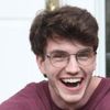 A young man with curly brown hair and glasses is smiling widely while wearing a maroon shirt.