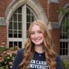 A young woman with long wavy blonde hair smiles while standing in front of a brick building with arched windows.