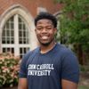A young man wearing a navy blue John Carroll University shirt smiles in front of a brick building with arched windows and