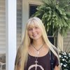 A young woman with long blonde hair smiles while standing outside near a white pillar and green plants.
