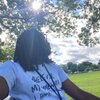 A person with dreadlocks and glasses sits under a tree on a sunny day, looking to the right with a grassy field behind.