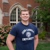 A young man wearing a navy blue John Carroll University t-shirt smiles standing outside in front of a brick building with