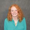 A young person with curly red hair smiles wearing a teal button-up shirt and two gold necklaces against a gray backdrop.