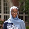 A smiling person wearing glasses and a light blue hijab stands outdoors in front of a building with arched windows.