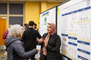 A woman wearing a pink hijab explains her research poster to an older woman holding a pencil and paper.