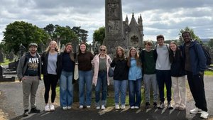 A group of ten people stand side by side in front of a large ornate stone cross in a cemetery under a cloudy sky.