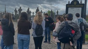 A group of people stand on a path listening to a man speaking near a Milltown Cemetery sign under a cloudy sky.
