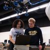 * Image content: Two students (one female with curly hair, one male with blonde hair/glasses) looking at a laptop screen