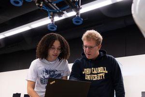 * Image content: Two students (one female with curly hair, one male with blonde hair/glasses) looking at a laptop screen