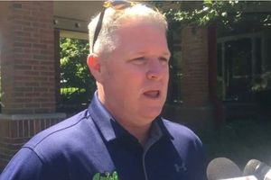 A man with short gray hair wearing a navy blue shirt speaks into microphones outside near a brick building and trees.