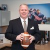 A man in a dark pinstripe suit smiles while holding a football in an office with sports photos and books.