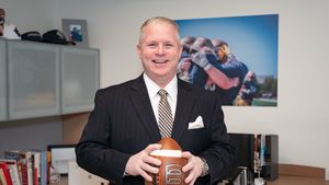 A man in a dark pinstripe suit smiles while holding a football in an office with sports photos and books.