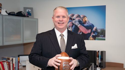 A man in a dark pinstripe suit smiles while holding a football in an office with sports photos and books.