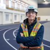 A young man wearing a white hard hat, safety glasses, and a reflective vest stands with arms crossed on an indoor running.