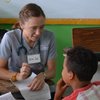 A woman wearing scrubs and a stethoscope talks to a boy sitting at a desk while holding a paper.