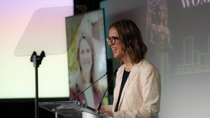 A woman wearing glasses and a light checkered blazer smiles while speaking at a clear podium with microphones.