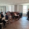 A man in a suit stands speaking to a seated audience of young adults in a bright room with large windows and flags.