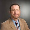 A man with short curly hair and a beard wears a tan blazer, blue shirt, and striped tie against a gray background.
