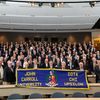 Dozens of smiling alumni pose together in a lecture hall, holding a blue banner that reads "John Carroll University Iota Chi
