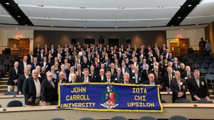 Dozens of smiling alumni pose together in a lecture hall, holding a blue banner that reads "John Carroll University Iota Chi