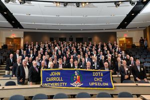 Dozens of smiling alumni pose together in a lecture hall, holding a blue banner that reads "John Carroll University Iota Chi