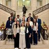 A group of twelve people wearing conference badges pose on a grand staircase with a statue and chandelier behind them.