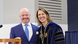 Two smiling individuals, a man in a blue suit and a woman in dark blue academic regalia with a gold chain, stand by a podium.