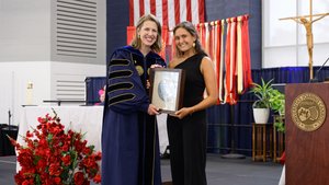 A smiling faculty member in blue academic regalia presents a framed silver medallion to a smiling student.