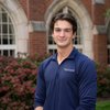 A young man wearing a navy blue John Carroll University pullover stands smiling in front of a brick building with arched.