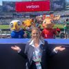 In the image, a woman is standing in front of a group of mascots, including a moose, eagle, and raccoon, at a sports event.