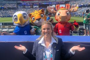 In the image, a woman is standing in front of a group of mascots, including a moose, eagle, and raccoon, at a sports event.