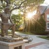 A bronze statue of a kneeling man holding a long object is in front of a brick building with sunlight shining through trees.