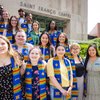 A diverse group of smiling students wearing blue and yellow stoles stand outside Saint Francis Chapel on a sunny day.