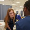 A woman in blue scrubs and gloves holds a penlight close to a man's face in a clinical setting.