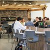 A group of students sit and eat at tables under a wooden pergola in a modern indoor dining area.