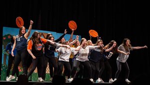 A group of young women on stage energetically pose with raised arms and hold basketball props against a painted backdrop.