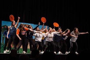 A group of young women on stage energetically pose with raised arms and hold basketball props against a painted backdrop.