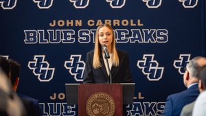 A young woman in a black blazer speaks at a podium with a microphone in front of a John Carroll Blue Streaks backdrop.