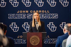 A young woman in a black blazer speaks at a podium with a microphone in front of a John Carroll Blue Streaks backdrop.