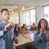 Two people write on a glass wall while three others watch and take notes inside a bright meeting room.