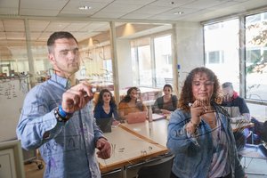 Two people write on a glass wall while three others watch and take notes inside a bright meeting room.