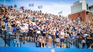 A large crowd of people stands and claps in stadium bleachers under a blue sky with flags waving.