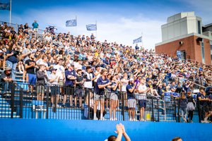 A large crowd of people stands and claps in stadium bleachers under a blue sky with flags waving.