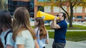 A man in a blue shirt speaks into a large yellow megaphone while three people stand nearby outside on a campus walkway.
